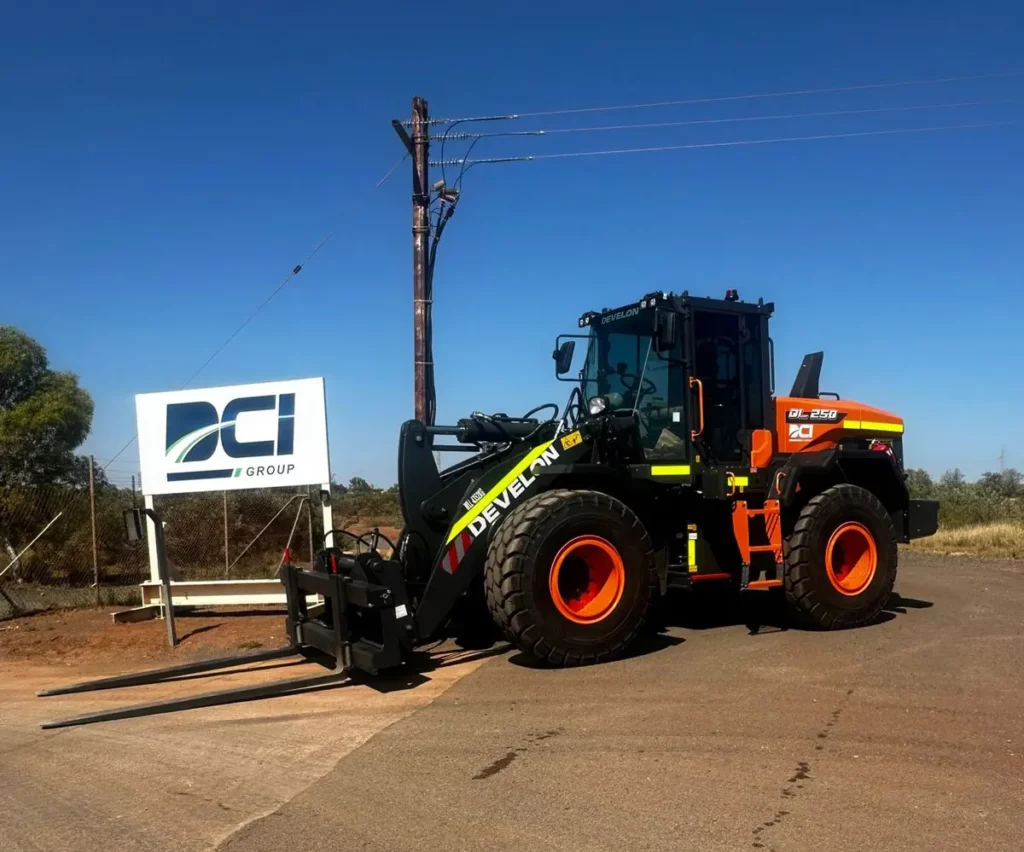 A large, orange and black forklift with "Develan" branding is parked on a road beside a white sign that reads "DCI Group" on a sunny day. The background features a fence, power pole, and clear blue sky.