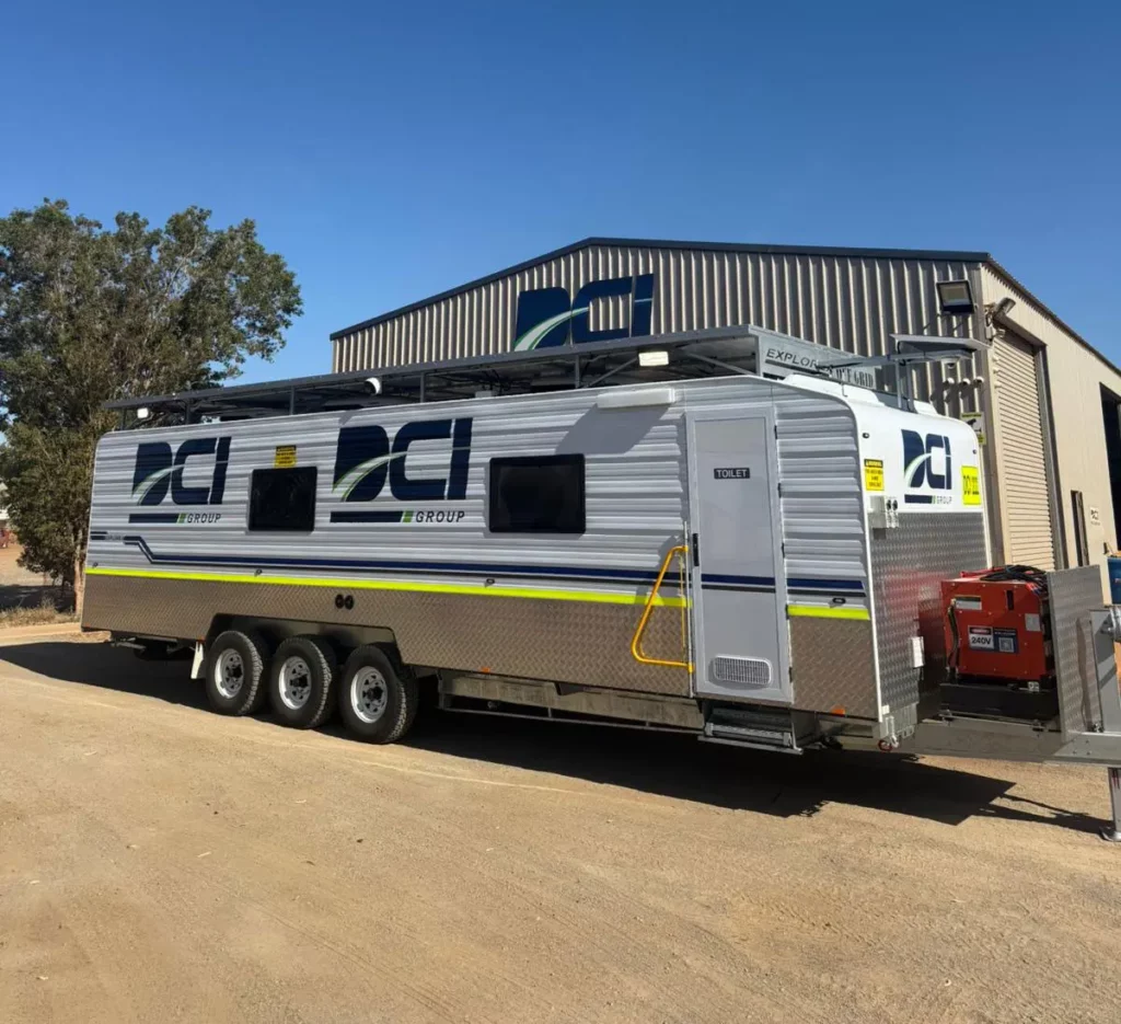 A large silver mobile trailer with triple axles and DCI Group branding is parked on a dirt lot in front of a metal building on a clear, sunny day. Trees and equipment are visible in the background.