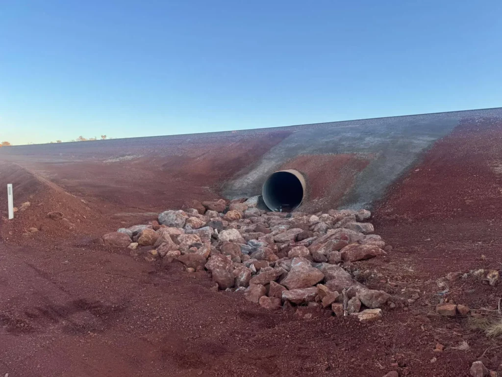A drainage pipe emerges from an embankment of reddish soil, with large rocks placed at its outlet under a clear blue sky—a typical scene in rail culvert remediation projects.