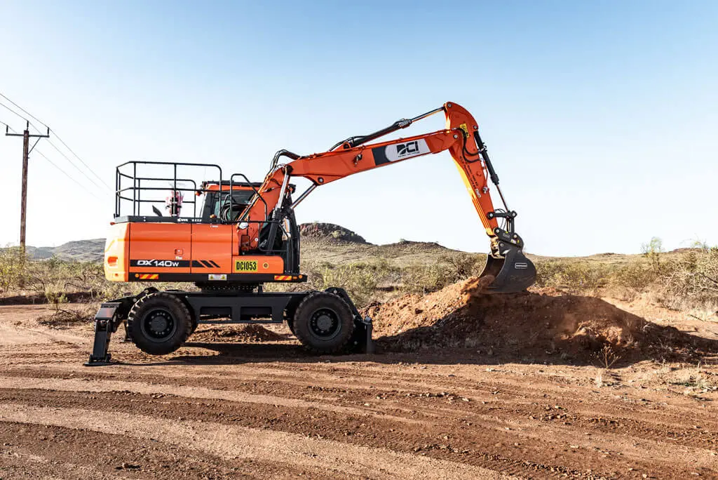 An orange wheeled excavator with a raised arm and bucket is digging into a mound of dirt on a construction site, with a clear sky and desert landscape in the background.