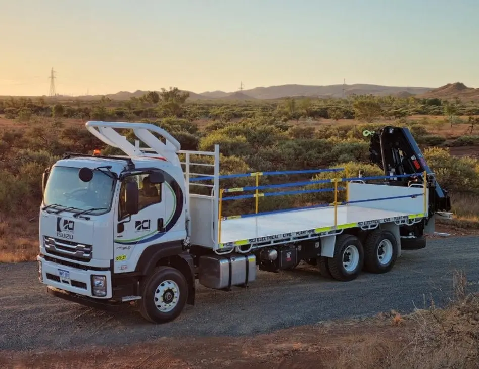 A white Isuzu flatbed truck with side rails and a mounted crane is parked on a dirt road in a rural area with shrubs and distant hills under a clear sky at sunset.