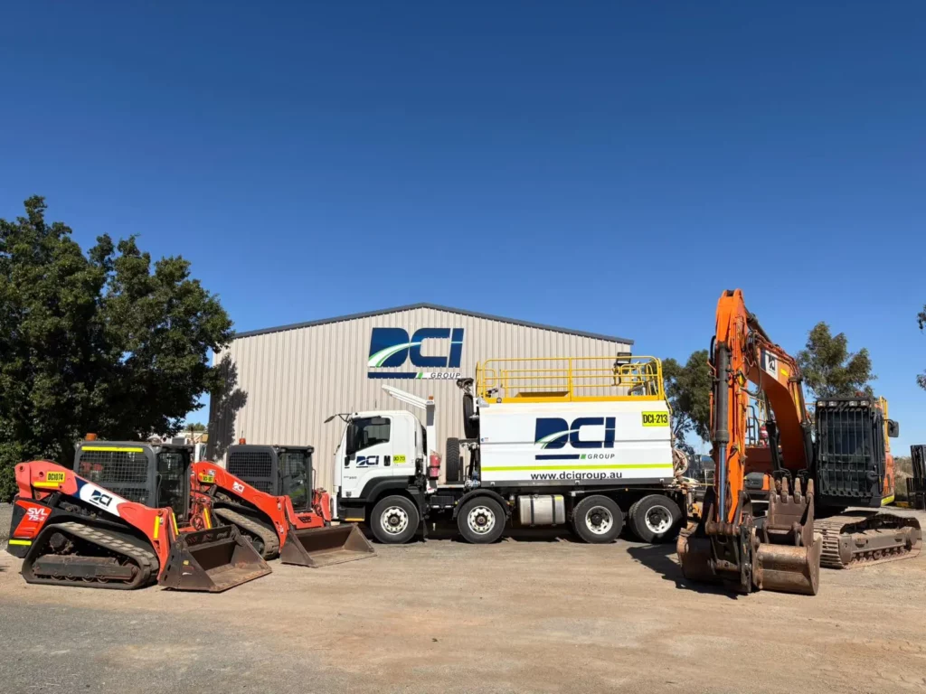 Four construction vehicles, including two loaders, a white truck with a yellow platform, and an orange excavator, are parked in front of a large industrial building with a "DCI" logo on it, under a clear blue sky.