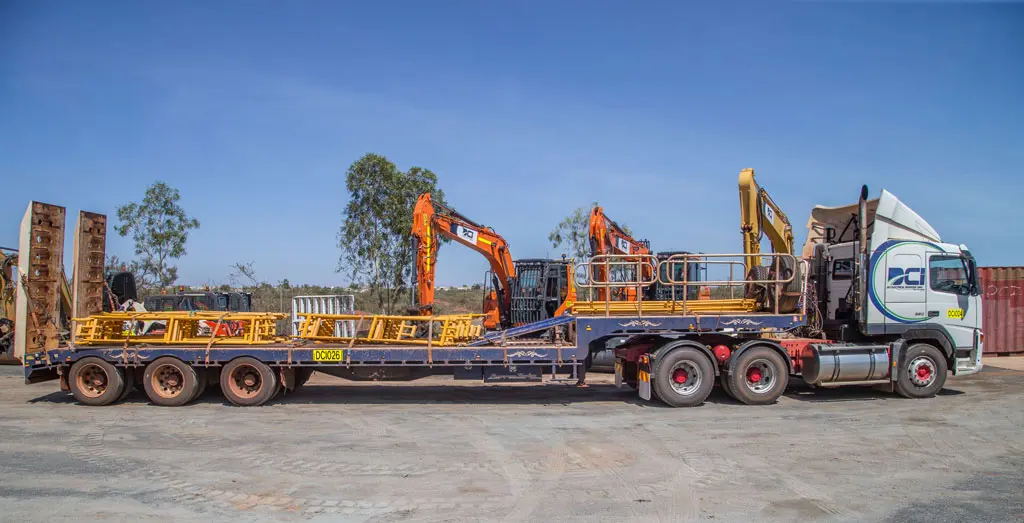 A large flatbed truck carries multiple yellow and orange excavators secured on its trailer, parked on a dirt lot with trees and clear blue sky in the background.