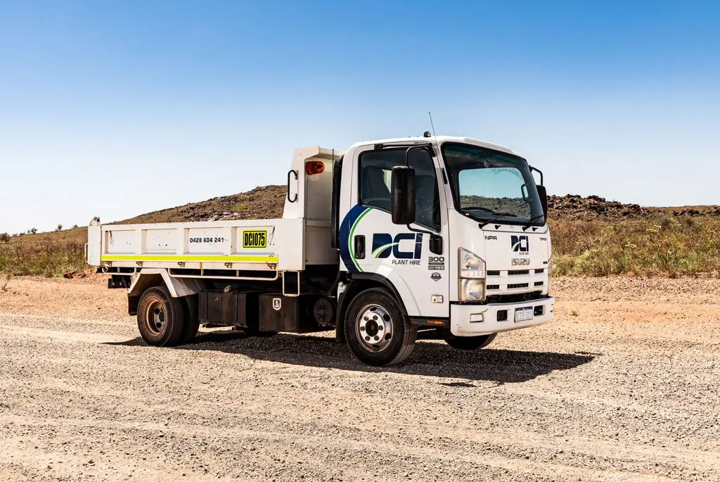 A white flatbed Isuzu truck with company logos is parked on a gravel road in a dry, open landscape under a clear blue sky.