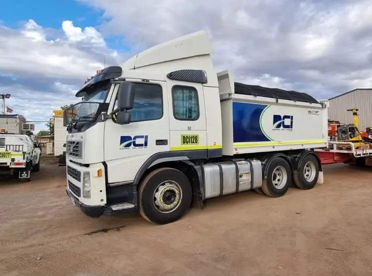 A white and blue dump truck with the DCI logo is parked on a dirt lot, with other vehicles and industrial equipment in the background under a cloudy sky.