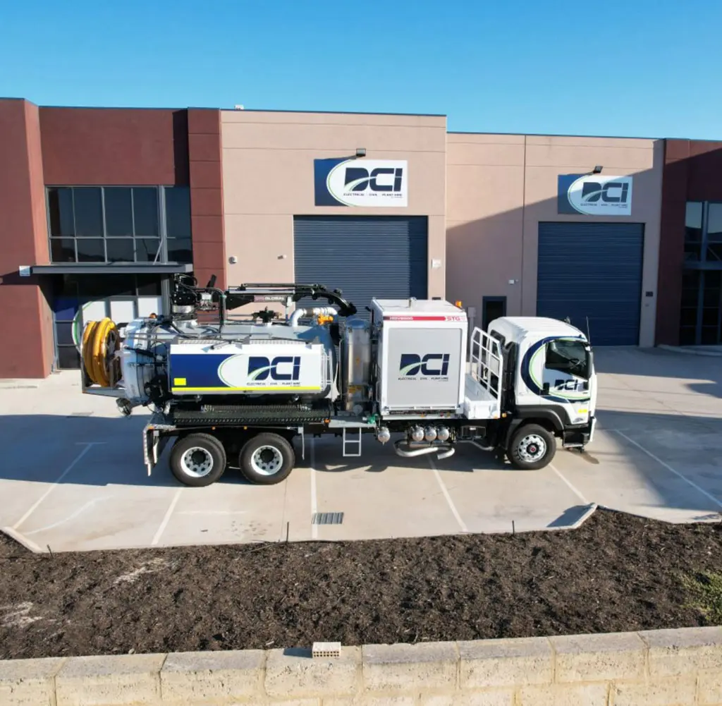A large white industrial vacuum truck is parked outside a modern building with "DCI" logos on both the truck and the building. The scene is bright and clear, with blue sky and closed garage doors.