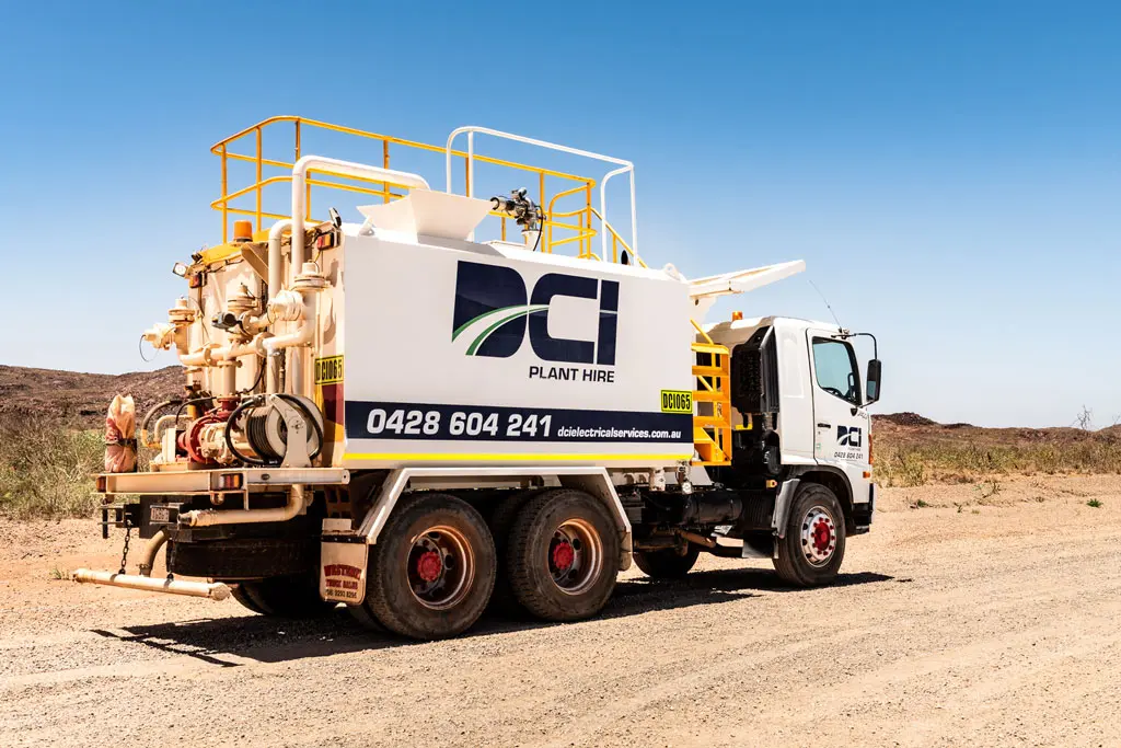 A white industrial truck with yellow railings and large machinery on the back is parked on a dirt road. The truck displays "Plant Hire" and a phone number on its side, with clear skies and dry terrain in the background.