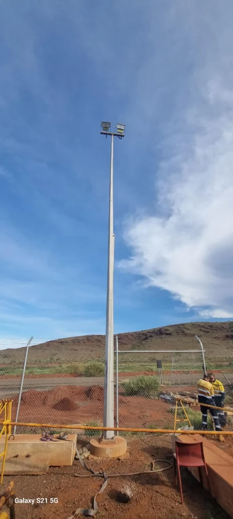 A tall floodlight pole stands near a wire fence, with a hill and partly cloudy sky in the background. A person in a yellow high-visibility shirt and helmet is working near the fence.